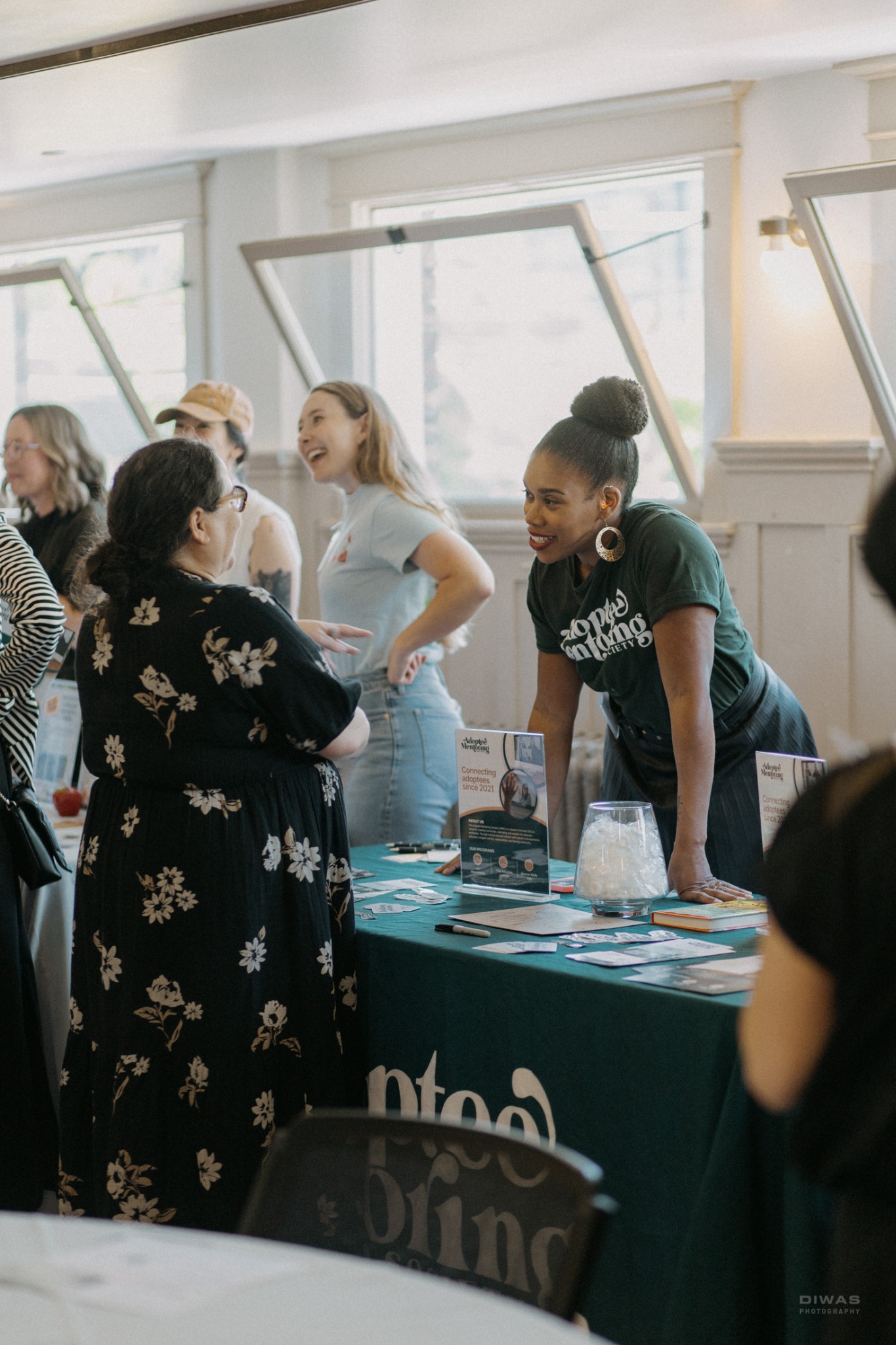 Angela Tucker from Adoptee Mentoring Society smiles while speaking with an attendee across an information table during the GrowFundMe: Thrive event. Other guests network in the background.