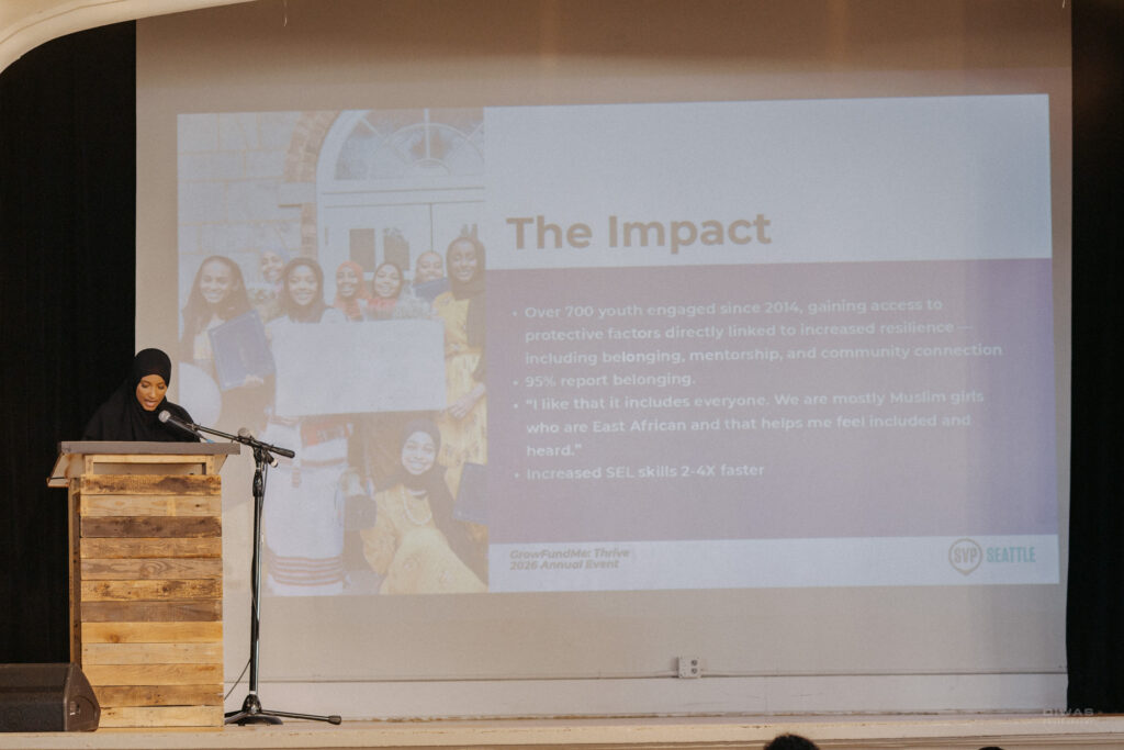 A nonprofit leader speaks from the podium during GrowFundMe: Thrive as a presentation slide highlights her organization's impact on youth and community belonging.