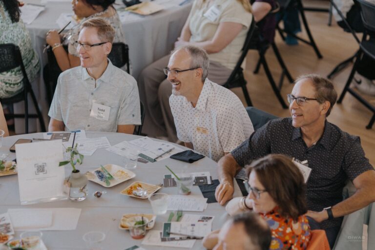 Three attendees smile and listen attentively during a GrowFundMe: Thrive presentation while seated together at a round table.
